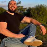 A smiling man in a black shirt and jeans sitting outdoors on a sunny day.