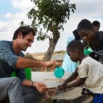 Man helping children wash hands outdoors with a water container.